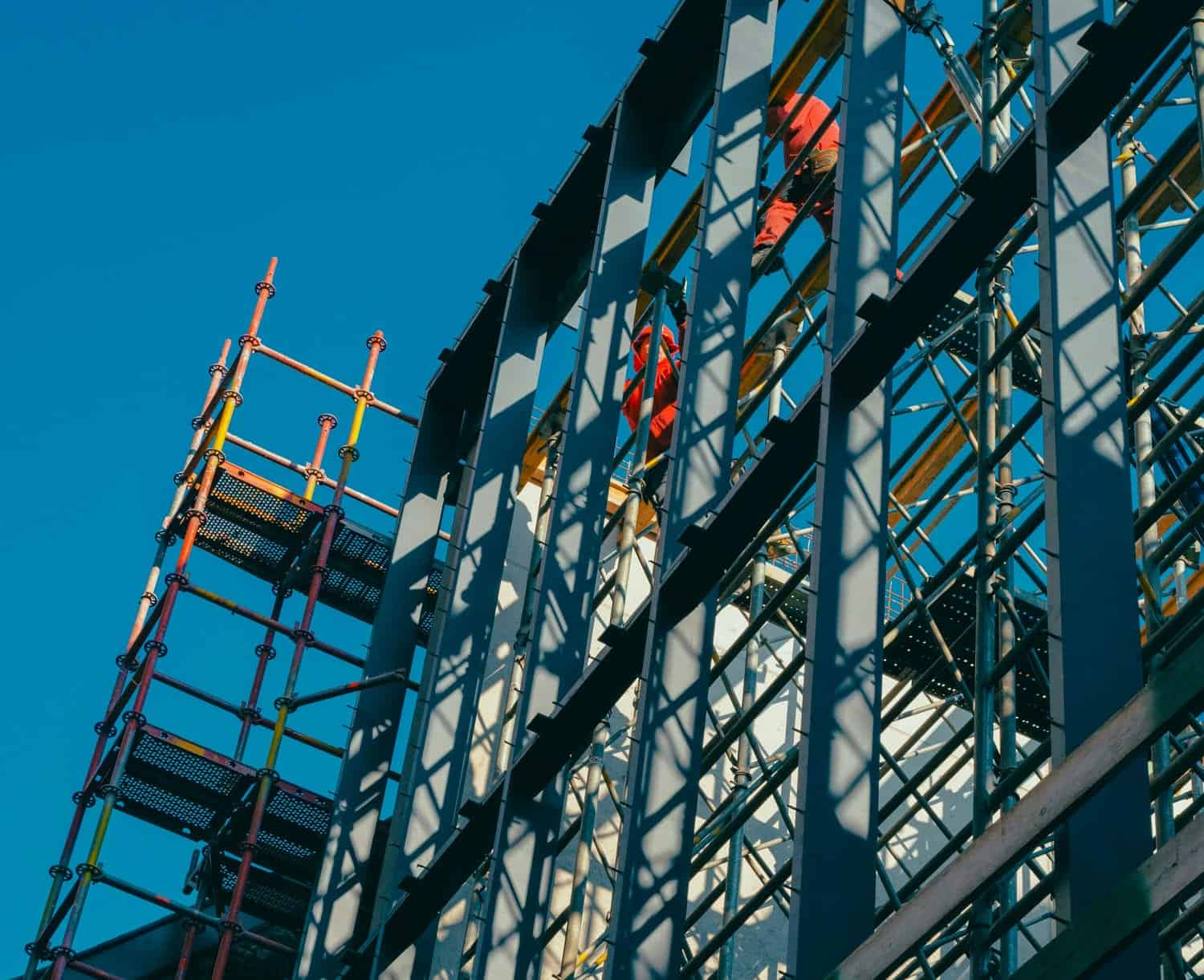 Industrial Construction Site With Steel Framework And Scaffolding Under A Clear Blue Sky, Showcasing Professional Building And Construction Services For Commercial And Residential Projects.